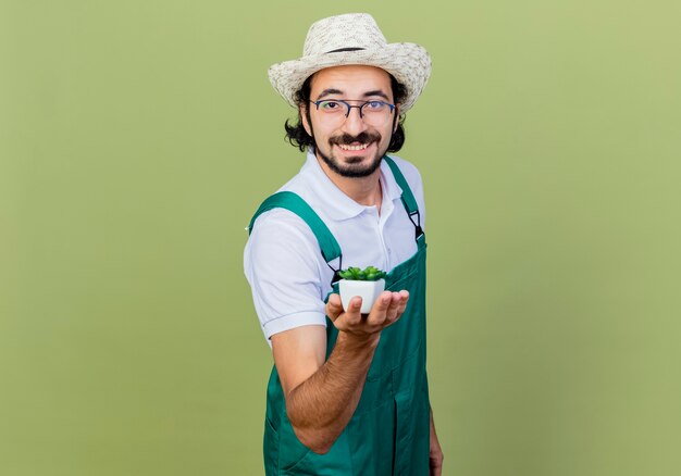 Jonge, gebaarde tuinmanmens die jumpsuit en hoed draagt die potplant toont die voorzijde glimlachend met gelukkig gezicht bekijkt die zich over lichtgroene muur bevindt