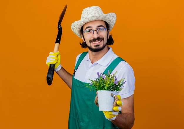 Jonge, gebaarde tuinmanmens die jumpsuit en hoed draagt die schop en potplant houdt die voorzijde glimlachend status over oranje muur bekijkt