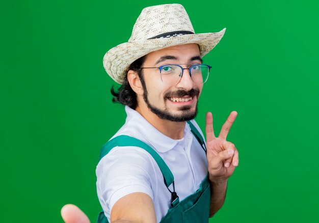 Jonge, gebaarde tuinmanmens die jumpsuit en hoed draagt ?? die selfie doet glimlachend toont v-teken dat zich over groene muur bevindt