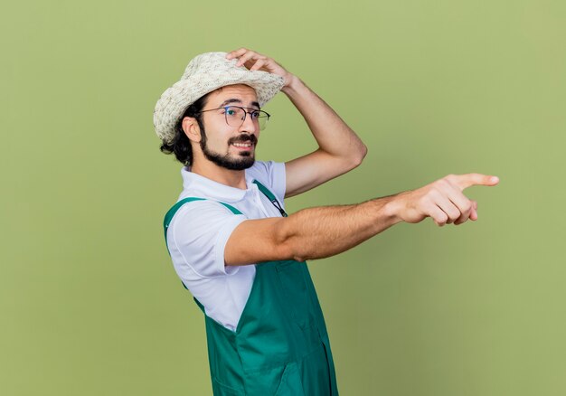 Jonge, gebaarde tuinmanmens die jumpsuit en hoed draagt wijst met wijsvinger naar iets dat zich over lichtgroene muur bevindt