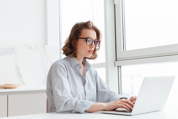 jonge geconcentreerde vrouw in gestreept overhemd met behulp van laptop tijdens het aanbrengen aan tafel in lichte appartement