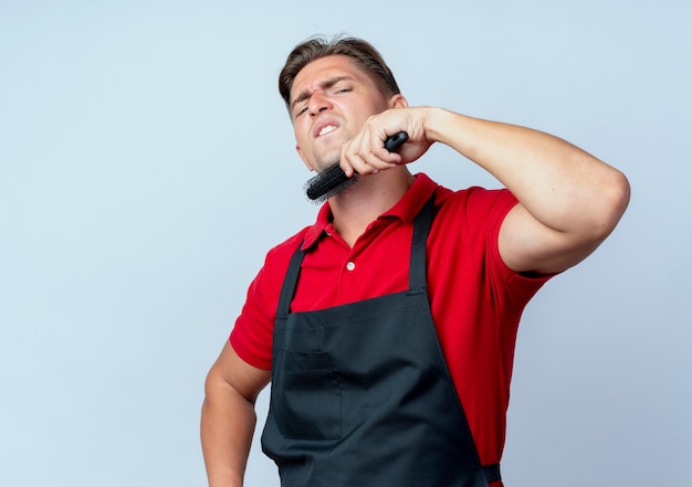 Jonge geërgerd blonde mannelijke kapper in uniform houdt kam op kin geïsoleerd op een witte ruimte met kopie ruimte