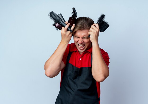 Jonge geërgerd blonde mannelijke kapper in uniform houdt kapper tools dicht bij het hoofd geïsoleerd op een witte ruimte met kopie ruimte