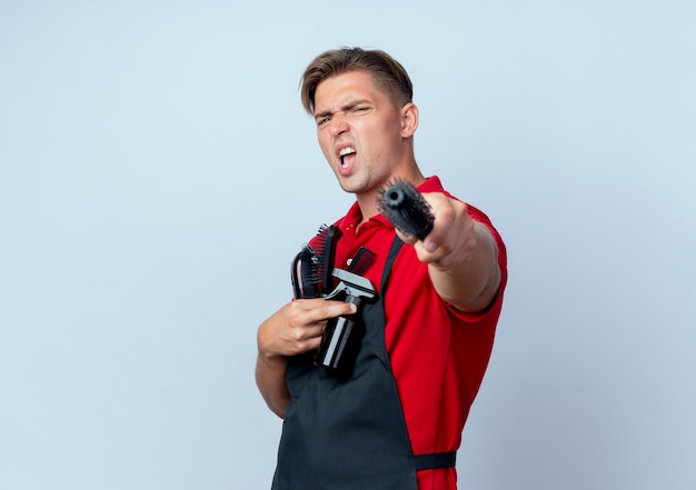 Jonge geërgerd blonde mannelijke kapper in uniform houdt kapper tools geïsoleerd op een witte ruimte met kopie ruimte