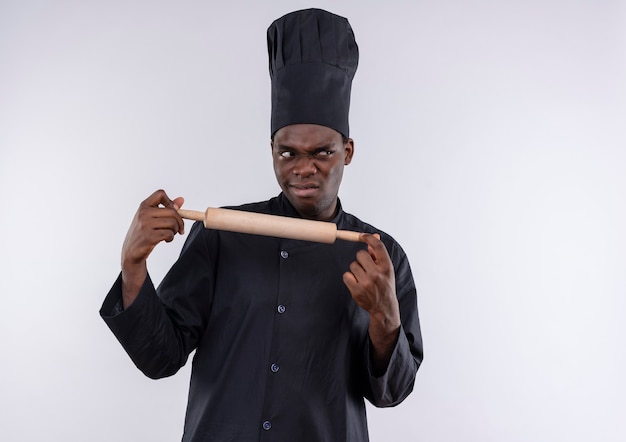 Jonge geïrriteerde Afro-Amerikaanse kok in uniform chef houdt deegroller nd kijkt naar kant op wit met kopie ruimte