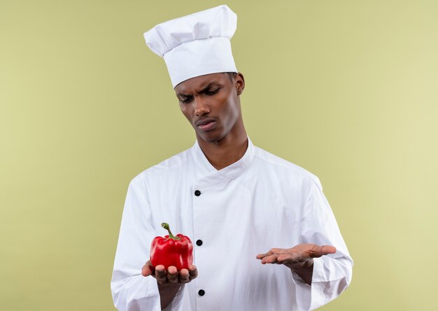 Jonge geïrriteerde Afro-Amerikaanse kok in uniform chef houdt rode peper en houdt de hand open geïsoleerd op groene achtergrond met kopie ruimte