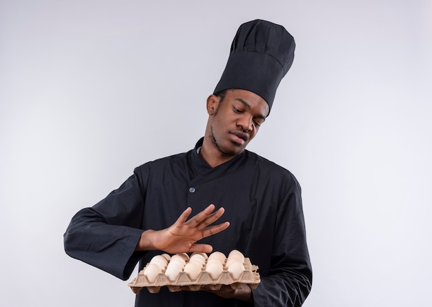 Jonge geïrriteerde afro-amerikaanse kok in uniform chef-kok houdt partij eieren en doet alsof hij wegduwt geïsoleerd op een witte achtergrond met kopie ruimte