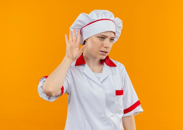 Jonge geïrriteerde blonde vrouwelijke chef-kok in uniforme gebaren van de chef kan geen teken horen dat op oranje muur wordt geïsoleerd