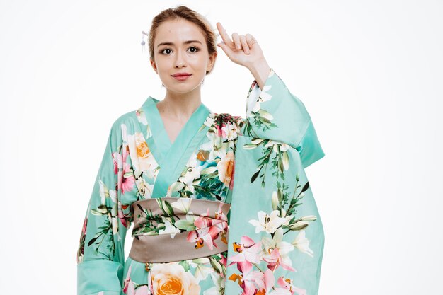 Jonge geisha vrouw in traditionele Japanse kimono kijkend naar de voorkant met een glimlach op een slim gezicht wijzend met de wijsvinger omhoog staand over de witte muur