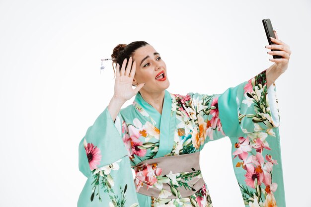 jonge geisha vrouw in traditionele japanse kimono met smartphone met een videogesprek blij en verrast glimlachend breed zwaaiend met een hand die over de witte muur staat