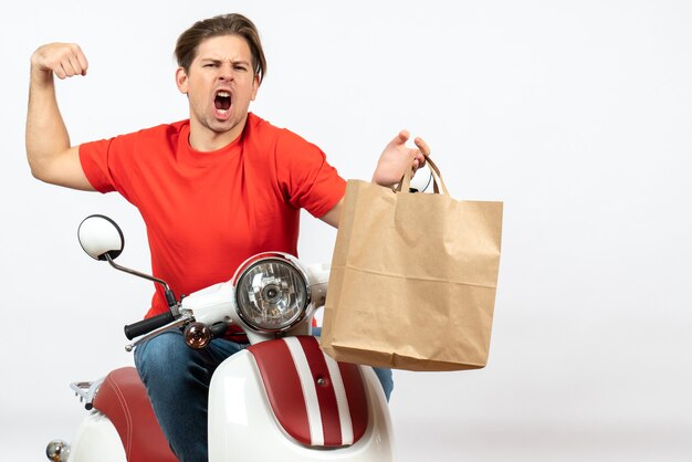 Jonge gekke emotionele koerierskerel in rood uniform zittend op de papieren zak van de scooterholding zijn gespierd op witte muur