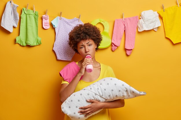 Jonge gekrulde moeder is moe van het zorgen voor pasgeboren, houdt baby gewikkeld in een deken, zuigt melk uit de fles, voelt liefde voor dochtertje, bezig met huishoudelijke taken en borstvoeding