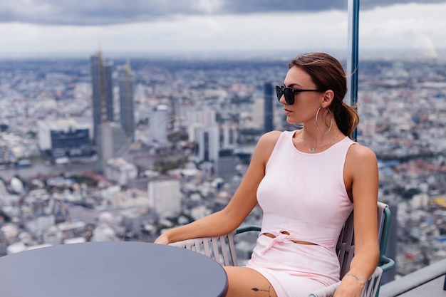 Jonge gelukkig lachend blanke vrouw reiziger in passende jurk en zonnebril op hoge verdieping in bangkok Stijlvolle vrouw verkennen geweldig uitzicht op de grote stad