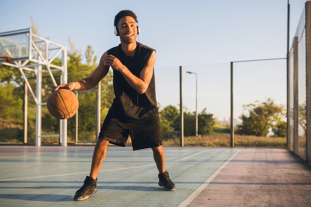Jonge gelukkig lachende man die sport doet, basketbal speelt bij zonsopgang, naar muziek luistert op een koptelefoon