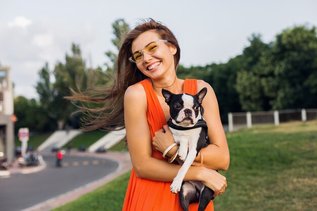 Jonge gelukkig lachende vrouw met boston Terriër hond in park, zonnige zomerdag, vrolijke stemming, spelen met huisdier, knuffels, oranje jurk, zonnebril, zomerstijl dragen