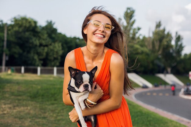 Jonge gelukkig lachende vrouw met boston Terriër hond in park, zonnige zomerdag, vrolijke stemming, spelen met huisdier, knuffels, oranje jurk, zonnebril, zomerstijl dragen