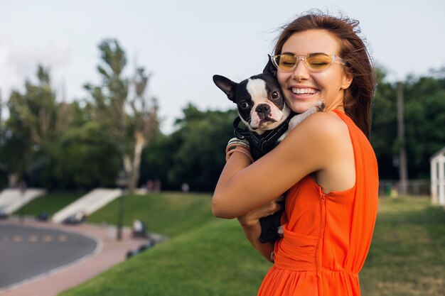 Jonge gelukkig lachende vrouw met boston Terriër hond in park, zonnige zomerdag, vrolijke stemming, spelen met huisdier, knuffels, oranje jurk, zonnebril, zomerstijl dragen