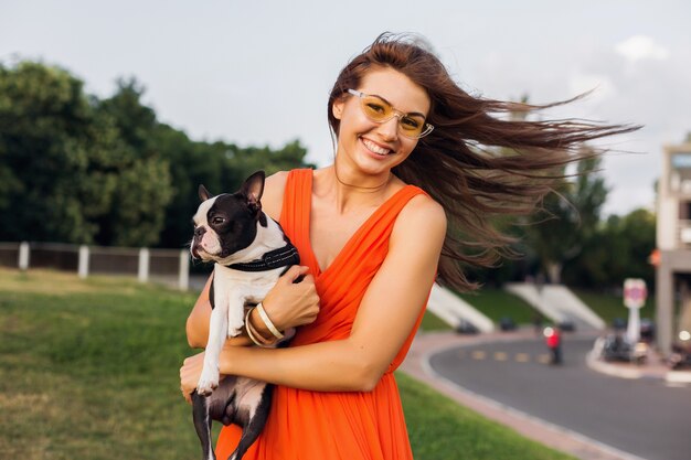 Jonge gelukkig lachende vrouw met boston terrrier hond in park, zonnige zomerdag, vrolijke stemming, spelen met huisdier, lang haar zwaaien, plezier maken, zomer modetrend