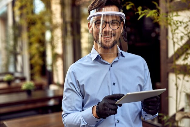 Jonge gelukkige café-eigenaar die vizier en beschermende handschoenen draagt tijdens het gebruik van een digitale tablet en naar de camera kijkt