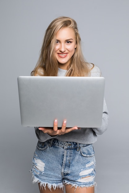 Jonge gelukkige glimlachende vrouw die in vrijetijdskleding laptop houdt en e-mail verzendt naar haar beste vriend die op grijze achtergrond wordt geïsoleerd