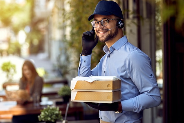 Jonge gelukkige koerier die een headset draagt tijdens het bezorgen van eten uit een restaurant