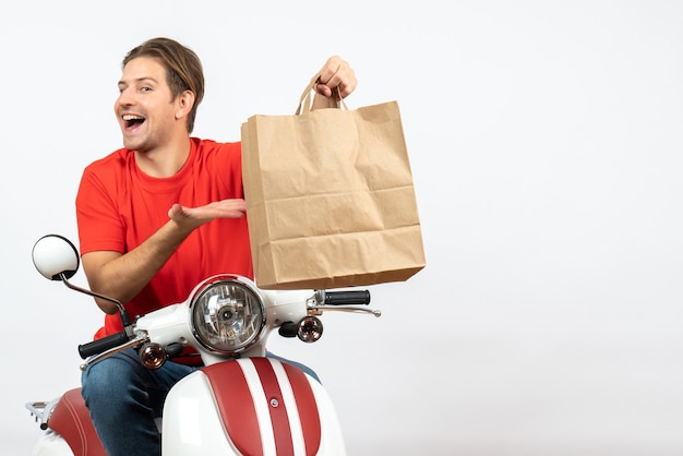 Jonge gelukkige koerierskerel in rood uniform zittend op de papieren zak van de scooterholding op witte muur