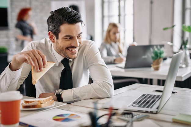 Jonge gelukkige manager die sandwich eet en computer gebruikt tijdens de lunchpauze op kantoor Er zijn mensen op de achtergrond