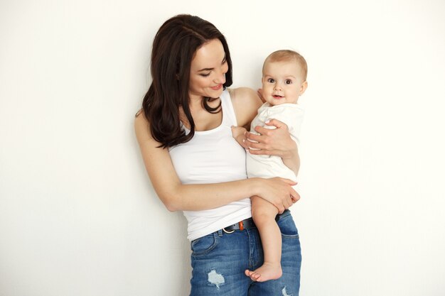 Jonge gelukkige moeder het glimlachen holding die haar babydochter bekijken over witte muur.