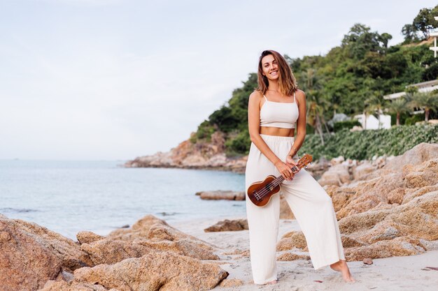 jonge gelukkige rustige blanke vrouw met ukelele op tropisch rotsachtig strand bij zonsondergang