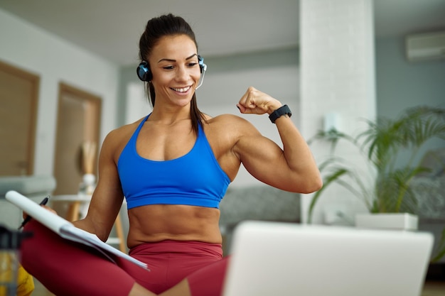 Jonge gelukkige sportvrouw die haar biceps buigt terwijl ze thuis een videogesprek voert via een laptop