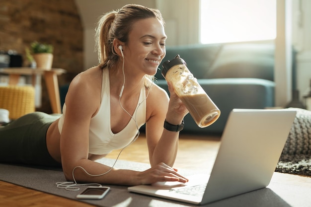 Jonge gelukkige sportvrouw die smoothie drinkt terwijl ze ontspant op de vloer en op het net surft op laptop