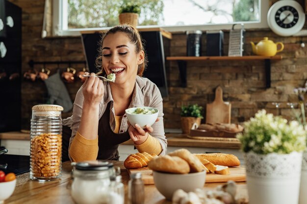 Jonge gelukkige vrouw die een gezonde maaltijd maakt en voedsel proeft in de keuken.