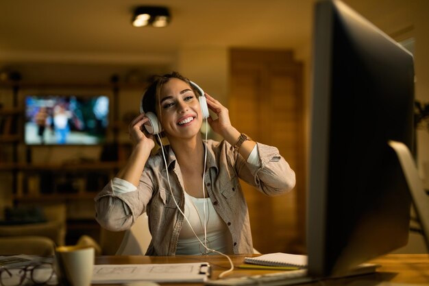 Jonge gelukkige vrouw die op het net surft op de computer terwijl ze 's avonds thuis naar muziek luistert op een koptelefoon