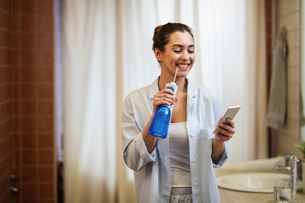 Jonge gelukkige vrouw die tandheelkundige waterflosser gebruikt en haar tanden schoonmaakt terwijl ze sms't op een mobiele telefoon in de badkamer