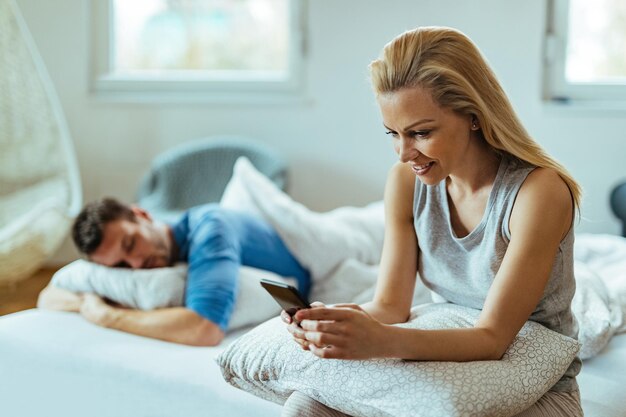 Jonge gelukkige vrouw met behulp van mobiele telefoon zittend op het bed. Haar vriendjes slapen achter haar.
