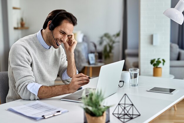 Jonge gelukkige zakenman die een hoofdtelefoon draagt terwijl hij thuis aan een laptop werkt