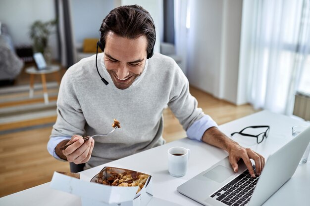 Jonge gelukkige zakenman die laptop gebruikt terwijl hij thuis een lunchpauze heeft