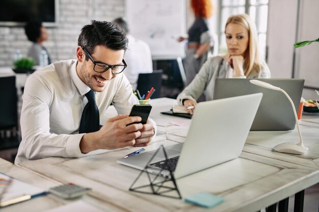 Jonge gelukkige zakenman die smartphone gebruikt en een bericht leest terwijl hij op kantoor werkt