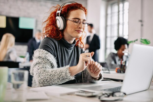 Jonge, gelukkige zakenvrouw die geniet van favoriete muziek via een koptelefoon terwijl ze op een computer op kantoor werkt. Er zijn mensen op de achtergrond