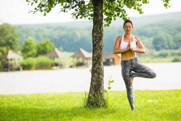 Jonge geschikte vrouw die yoga in Park dichtbij meer en boom doet