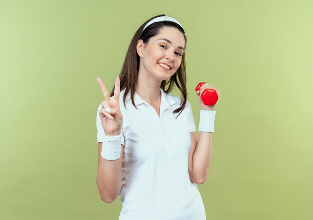 Jonge geschiktheidsvrouw in hoofdband die met domoor het glimlachen tonen die overwinningsteken tonen die zich over lichte muur bevinden