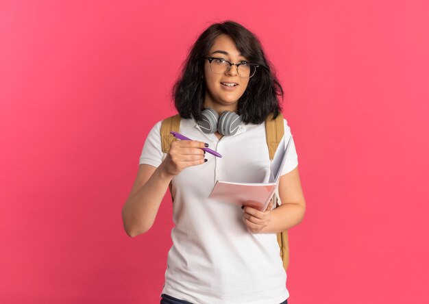 Jonge geschokt vrij Kaukasisch schoolmeisje met koptelefoon op nek bril en rugtas houdt pen en notitieblok op roze met kopie ruimte