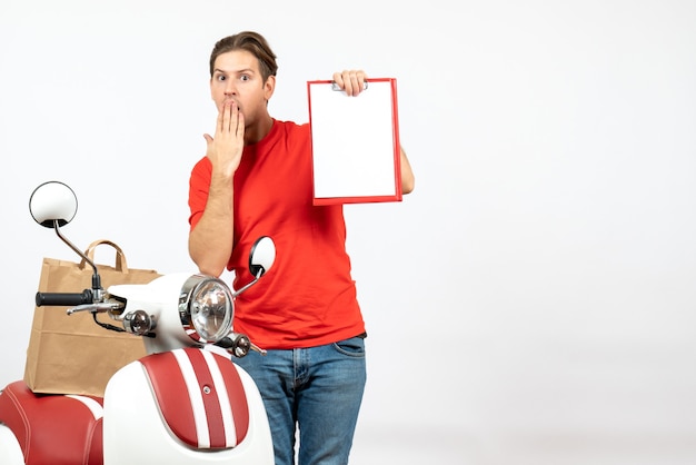 Jonge geschokte bezorger in rode uniform staande in de buurt van scooter met document op witte muur