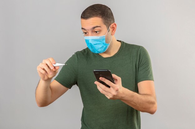 Jonge geschokte mens in medisch masker die digitale thermometer bekijken en een smartphone houden die ter beschikking op wit wordt geïsoleerd