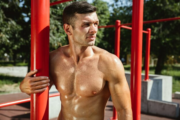 Jonge gespierde shirtless blanke man tijdens zijn training op horizontale balken op de speelplaats in zonnige zomerdag. Zijn lichaam buiten trainen. Concept van sport, gezonde levensstijl, welzijn.