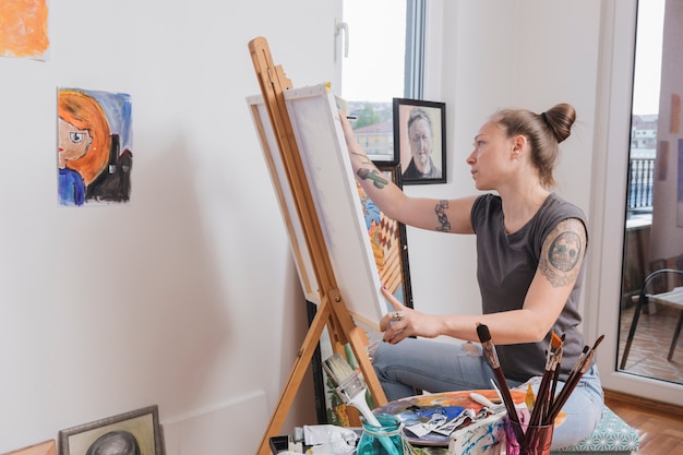 Gratis foto jonge getatoeëerde vrouw schilderij foto zitten in de kunststudio