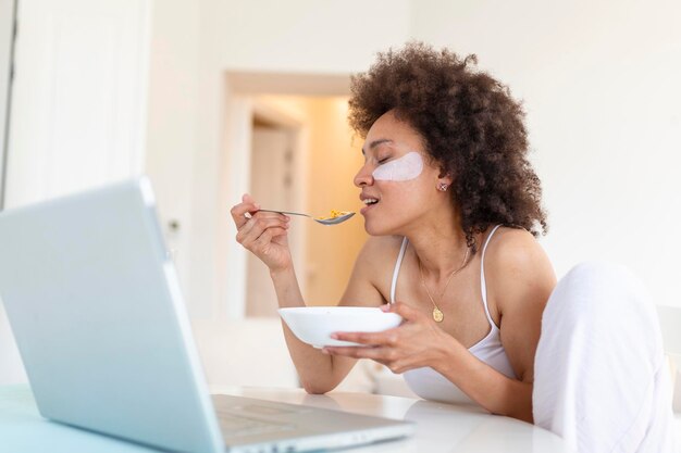 Jonge geweldige vrouw die binnen aan tafel zit met een laptop die cornflakes vasthoudt Kijkend naar een laptopcomputer en praat met haar vrienden via een videogesprek