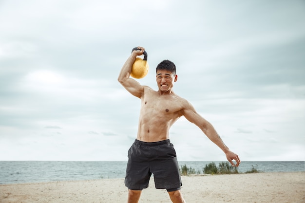 Jonge gezonde man atleet oefening met het gewicht op het strand. Signle shirtless training voor mannelijk model aan de rivierzijde op zonnige dag. Concept van een gezonde levensstijl, sport, fitness, bodybuilding.