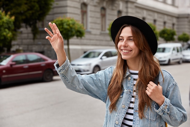 Jonge glimlachende jonge Europese vrouw loopt buiten, zwaait met de hand als vriend aankondigt in de verte