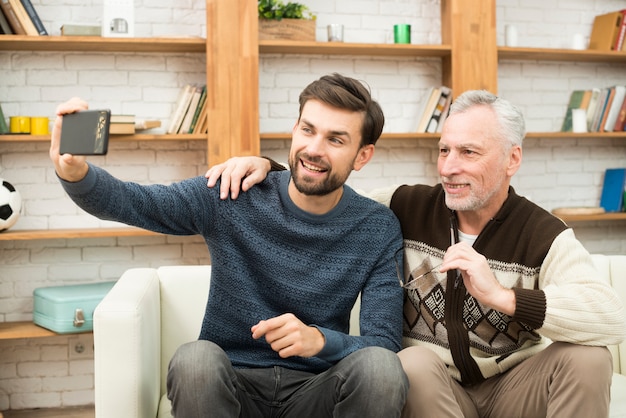 Jonge glimlachende kerel en de oude mens die selfie op smartphone op sofa nemen